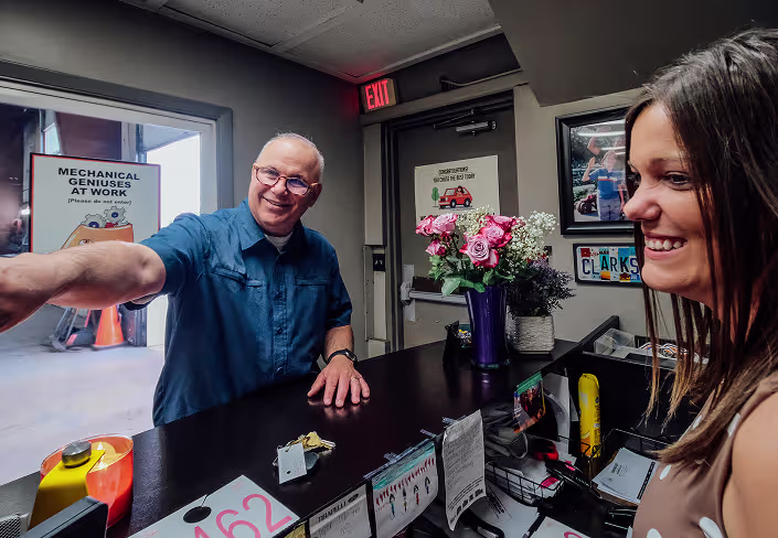 Auto repair in Naperville, IL at Clark's Car Care. A smiling man points while talking to a woman at a counter with flowers, keys, and papers. The background features posters, a door, and a Mechanical Geniuses At Work sign.
