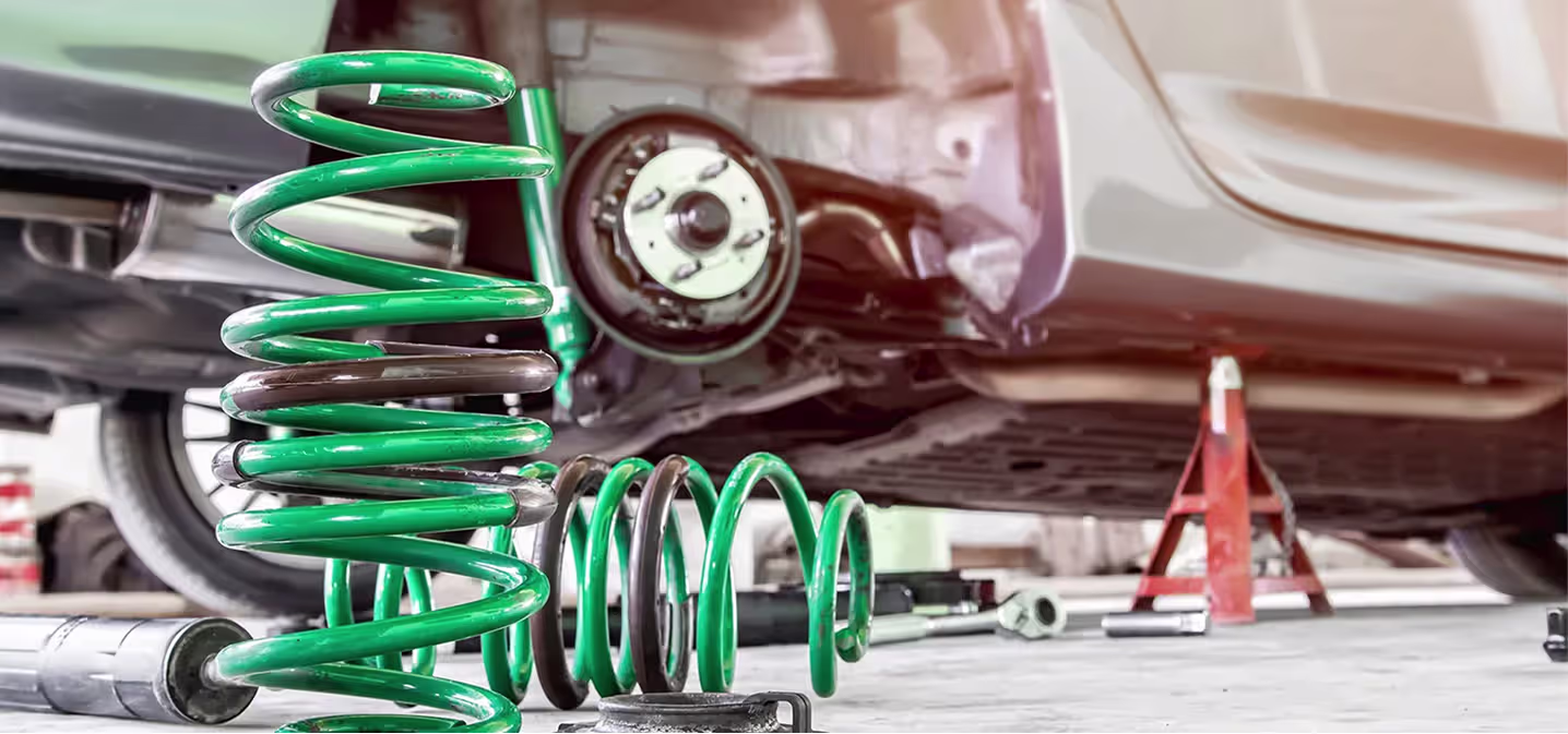 Steering & suspension repair in Naperville, IL at Clark's Car Care. A close-up of green coil springs on the floor next to a car elevated on red jack stands, with the cars rear wheel removed and tools scattered around.