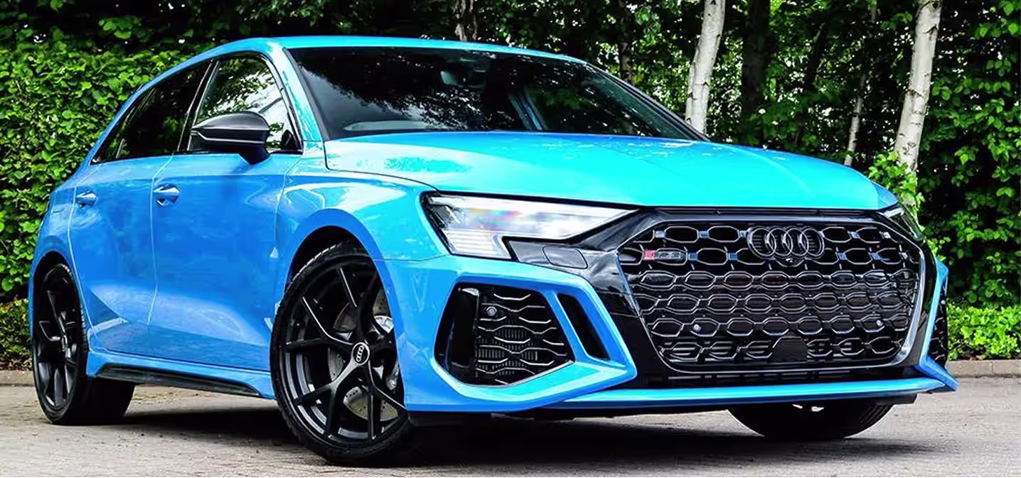 European vehicle repair in Naperville, IL at Clark's Car Care. A bright blue Audi hatchback with black alloy wheels is parked on a paved area, surrounded by green trees in the background.