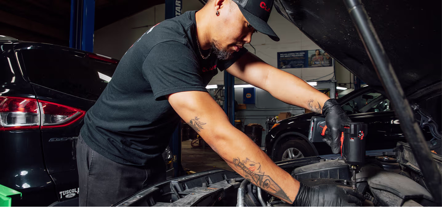 Engine repair and replacement in Naperville, IL at Clark's Car Care. A mechanic with tattoos and black gloves uses a power tool to work on a car engine in an auto repair shop, with other vehicles in the background.