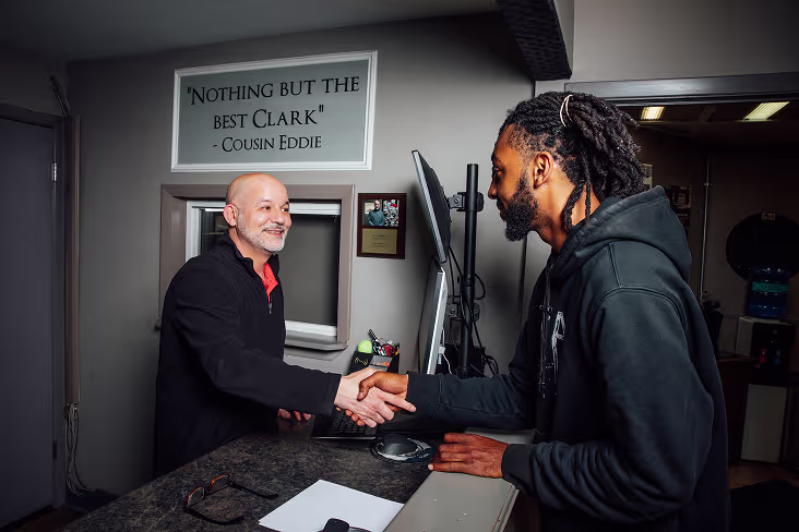 Two men shake hands across a counter in an office. One is bald, the other has dreadlocks. A sign on the wall behind them reads, Nothing but the best Clark—Cousin Eddie. A clipboard and computer are on the counter.