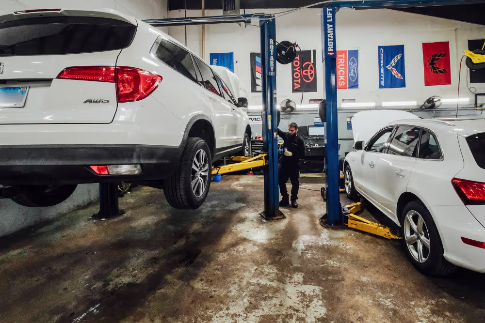 A mechanic works in an auto repair shop with two white SUVs on hydraulic lifts. Colorful banners hang on the wall in the background.