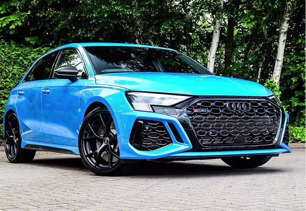 A bright blue Audi hatchback is parked on a paved area with green trees in the background. The car features black alloy wheels and a large honeycomb grille.