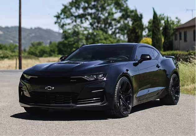 A black Chevrolet Camaro sports car is parked on a paved road with greenery and trees in the background under a clear sky.