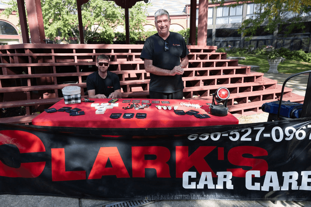 Community support by Clark's Car Care. Image of two team members behind a branded table displaying items at an outdoor event in Naperville, IL.
